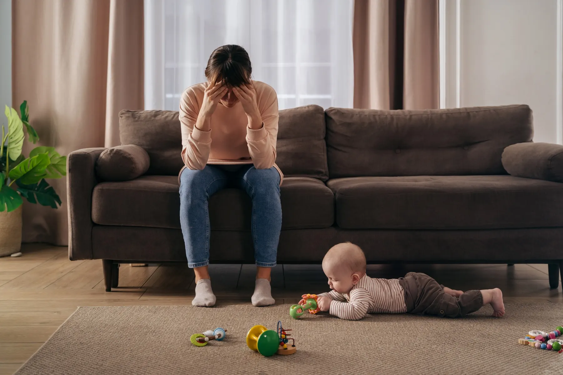 Une maman épuisée assise sur le canapé pendant que son bébé joue